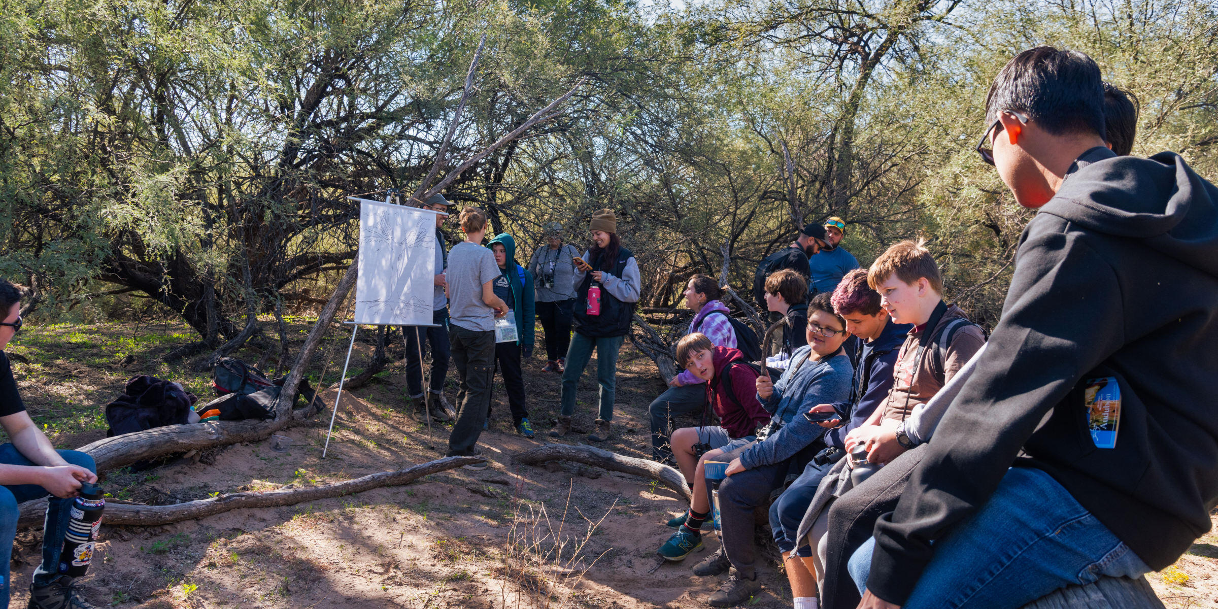 A group of high school students sit around an outdoor presentation under a canopy of mesquite trees.