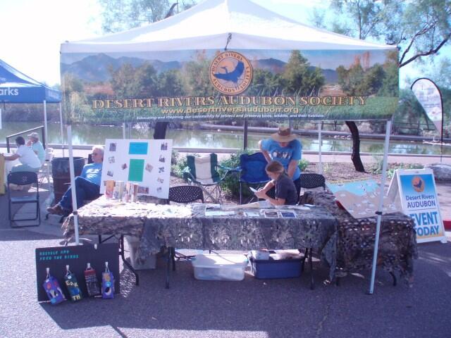 Volunteers in blue Desert Rivers Audubon shirts displaying Chapter education materials with a pond and mesquite trees as a backdrop.