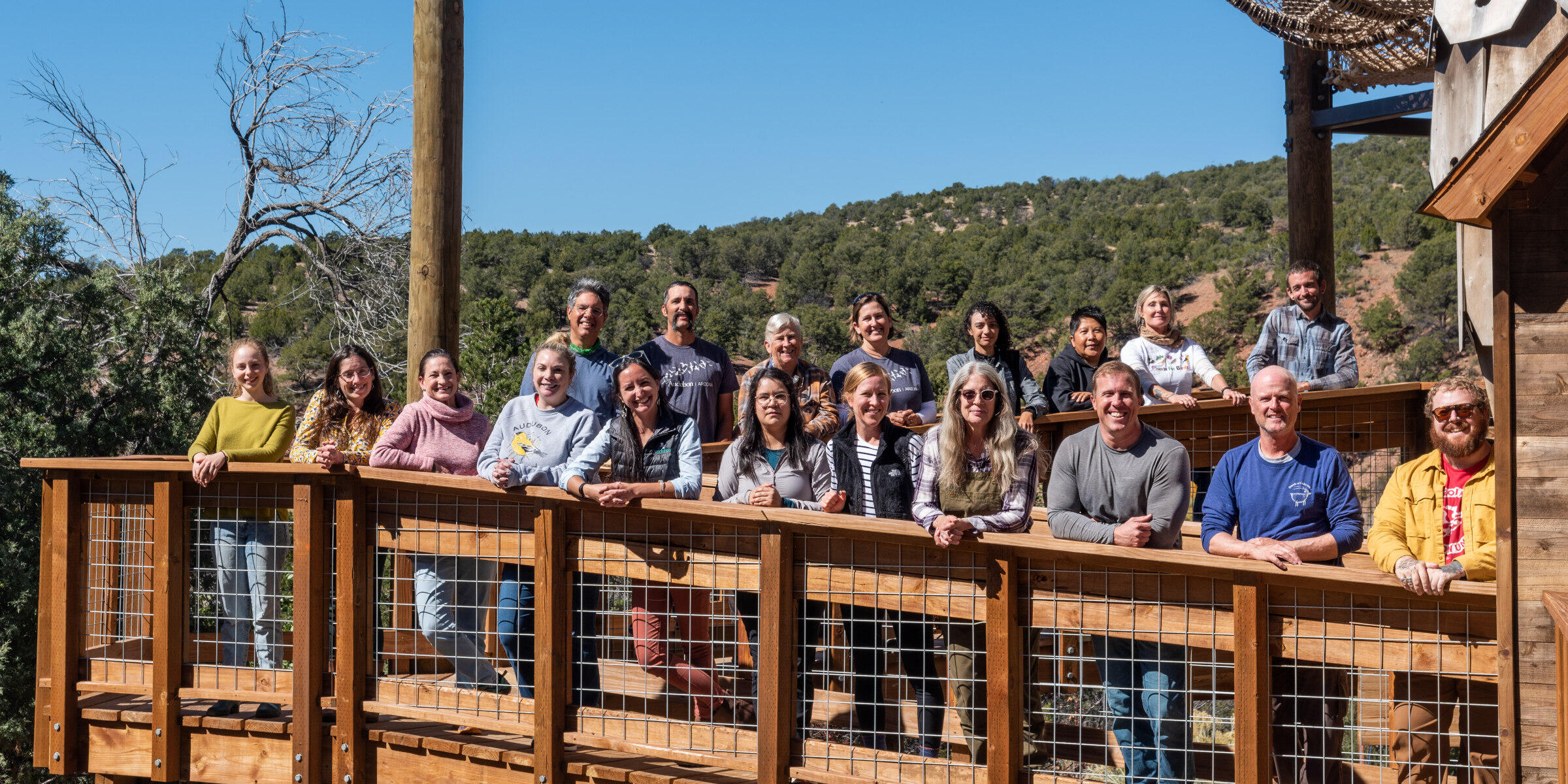 A group photo of nineteen adults standing on a wooden ramp to a playground with juniper forest in the background.