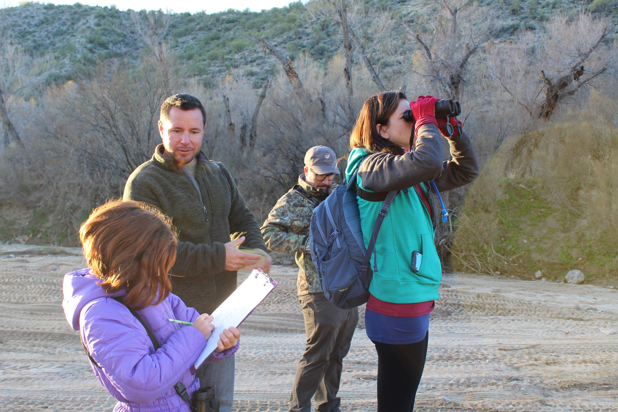 In a dry riverbed, a woman looks at a bird with binoculars while a little girl records the sighting on a piece of paper. Two men in the background get out their binoculars and field guide. It is sunny but also looks cold.