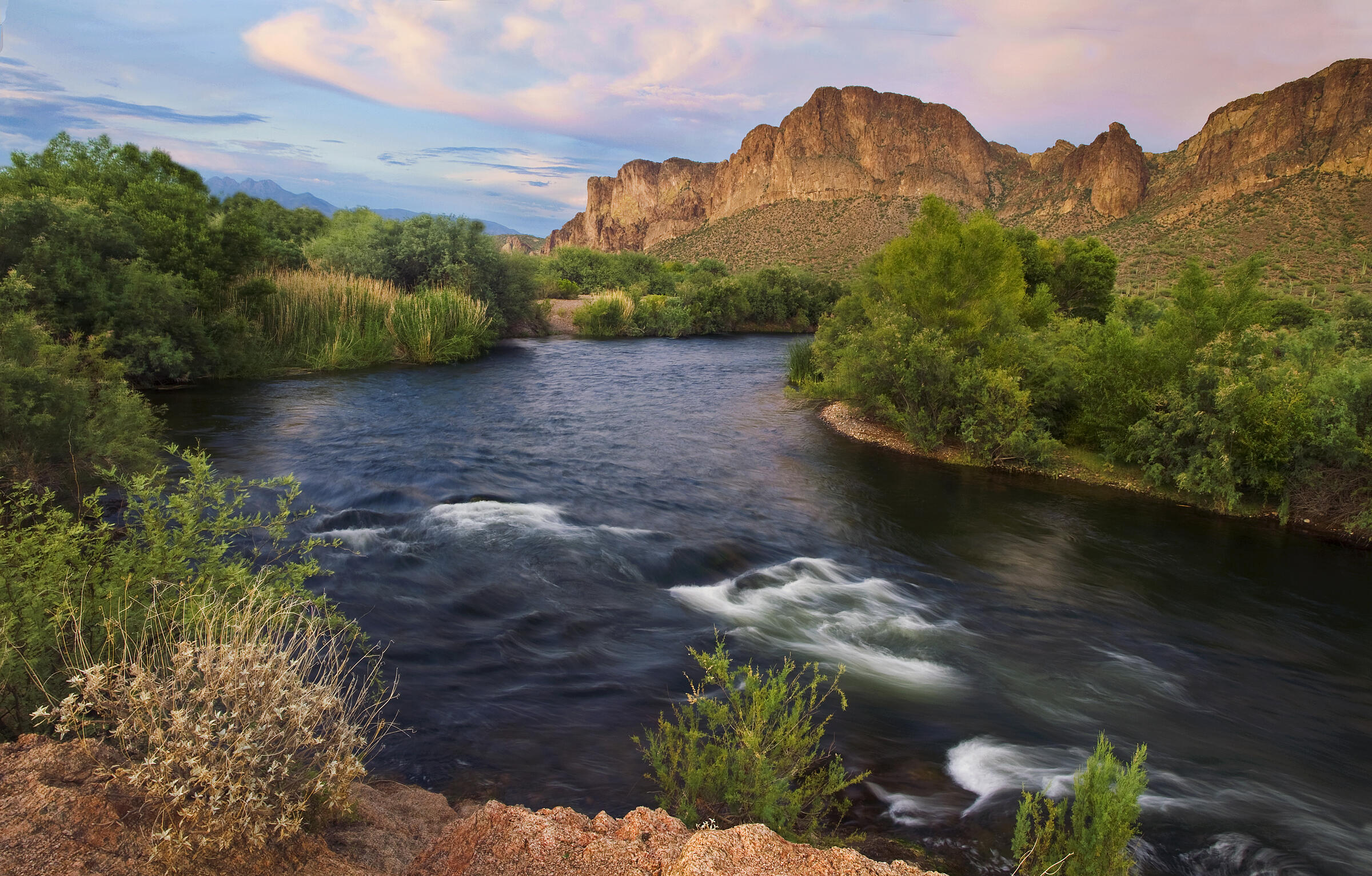 A slow exposure exaggerates the ripples of the deep blue Salt River against a backdrop of green trees, cattails, and the rugged cliff faces of the Goldfield Mountains.